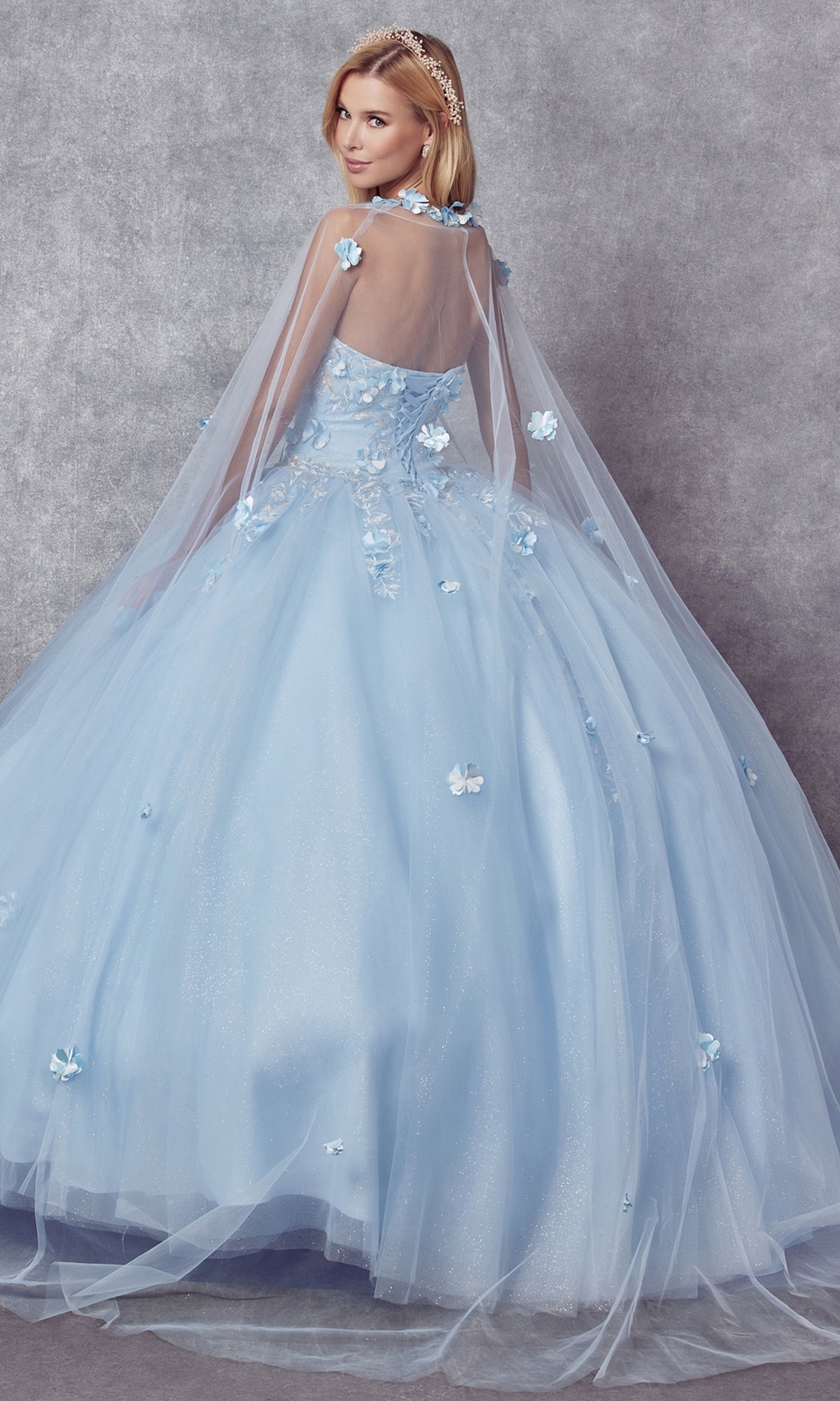 A woman poses against a gray textured backdrop, looking over her shoulder in the Juliet 1435 Strapless Ball Gown Quinceañera Dress, featuring light blue embroidery, floral appliqués, and a sheer cape.